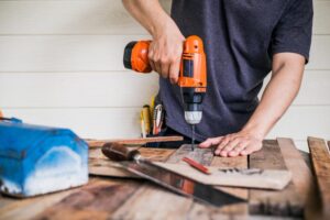 Un homme perce une planche de bois avec une perceuse orange, utilisant des outils de construction sur un établi rustique