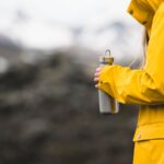 Une personne en veste jaune tient une gourde devant un décor montagneux